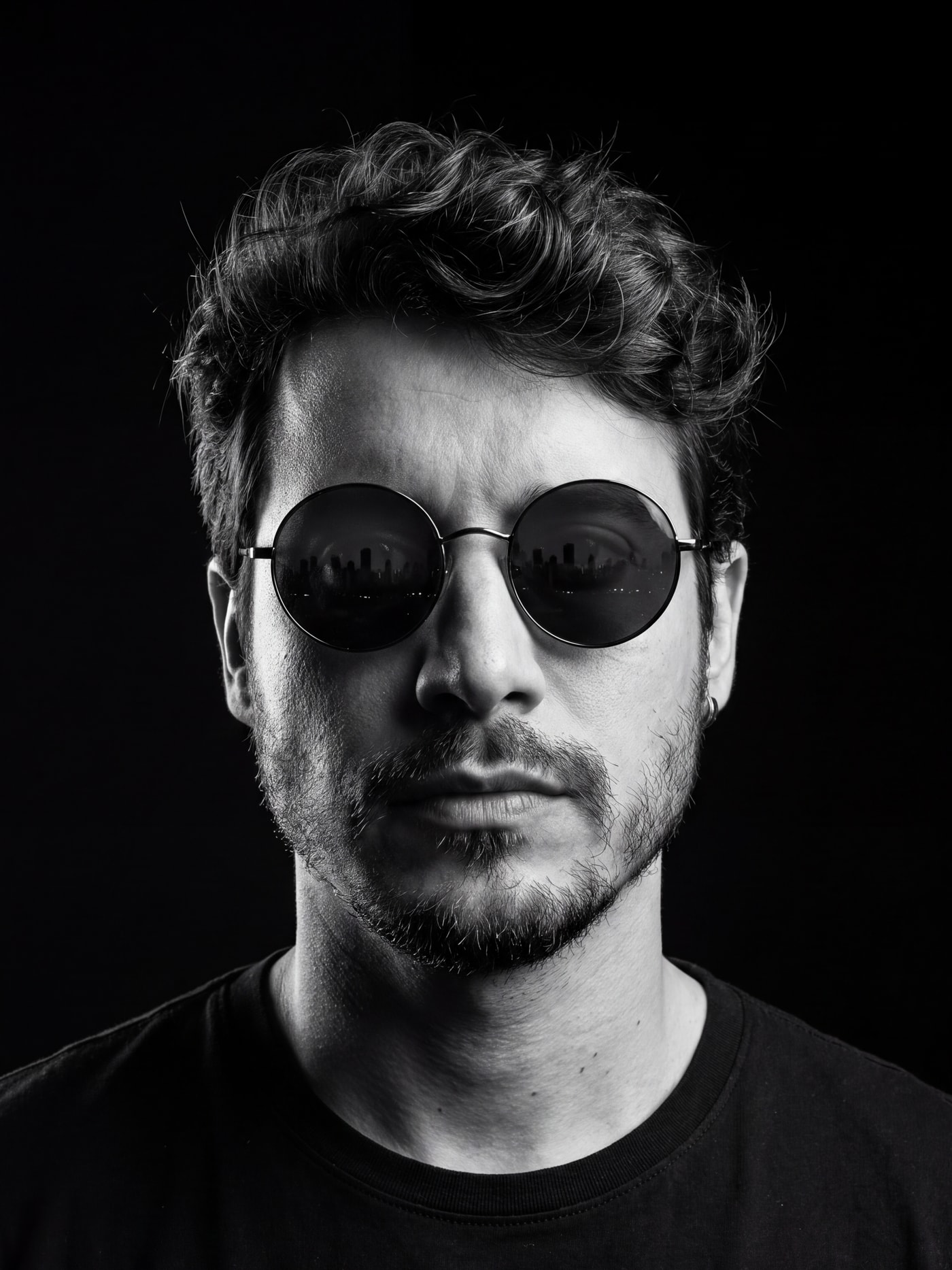 Retrato de James Olaya em preto e branco, de óculos escuros redondos, sobre fundo preto